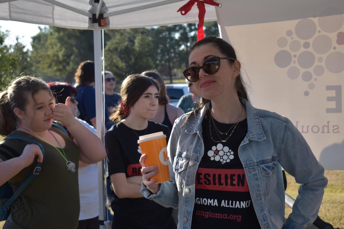 woman in a black shirt smiling with a yellow coffe cup and a group of people behind her