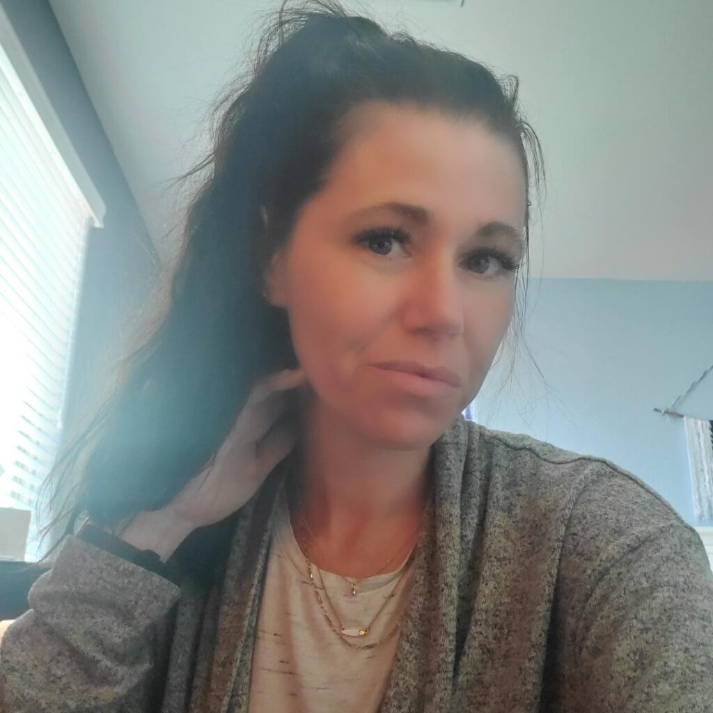 woman looking at a camera with a long brown ponytail and a grey sweater over a white tank top wearing a few gold necklaces.