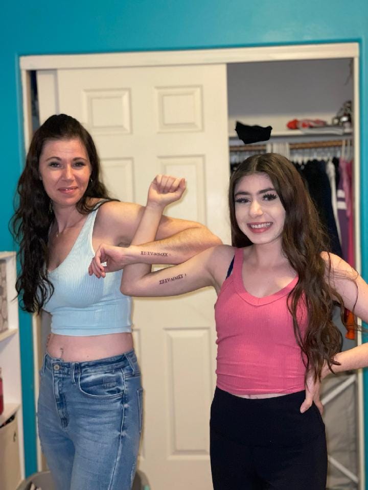 two girls with arms linked smiling, showing off Roman Numeral tattoos in a room with teal walls and an open closet behind them