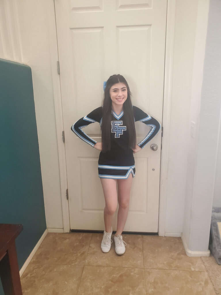 a girl in a cheer uniform standing in front of a door with her hands on her hips, smiling