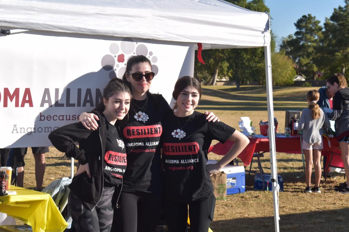 a woman and 2 teens standing in front of a large banner outside in black shirts and leggins. the shirts match and read "Angioma Alliance"