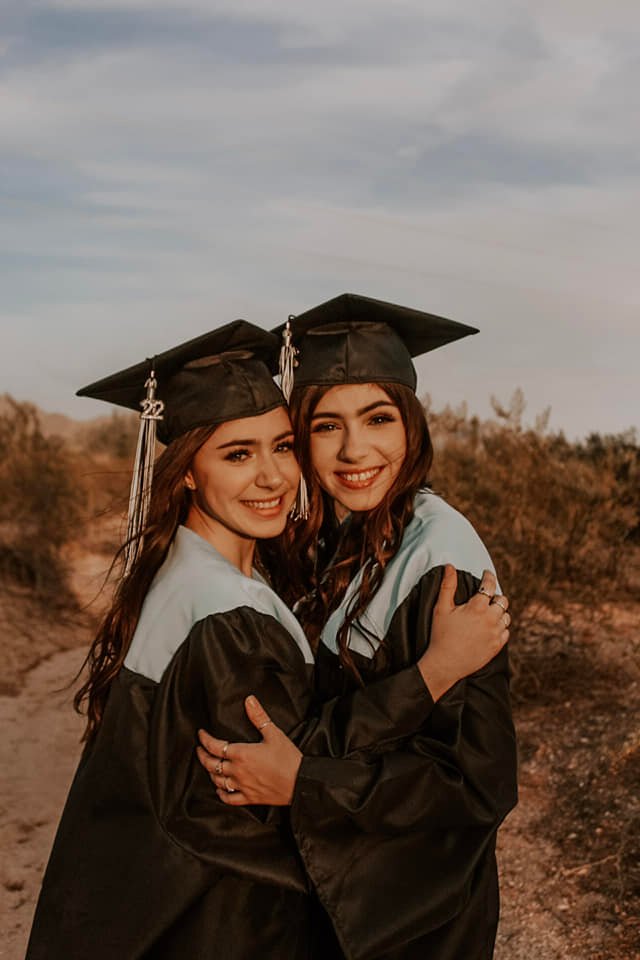 twin girls in matching black graduation caps with '22 tassels, with black gowns and light blue, hugging each other and smiling in the desert.