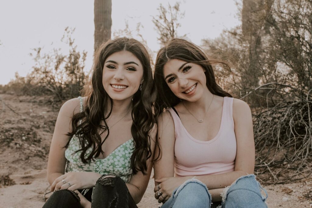 twins with long, dark hair sitting outside with cactus and brush behind them, both smiling at the camera