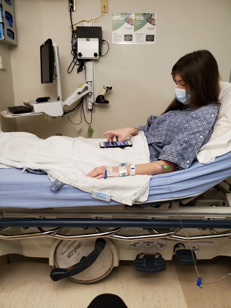 A young, dark-haired girl with an IV in a hospital bed wearing a gown and a mask, looking at her phone in her lap