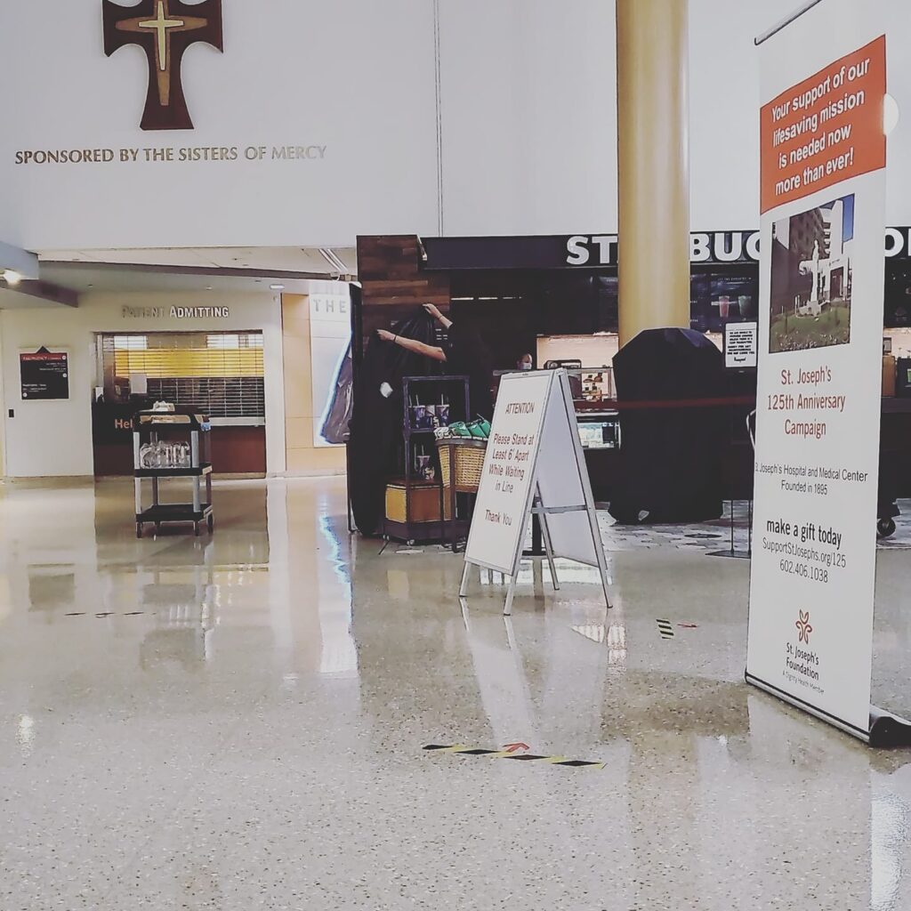 An empty hospital lobby with a cross and the words Sponsored by the sisters of mercy on t wall, Signs standing on the floor for St. Joseph's hospital