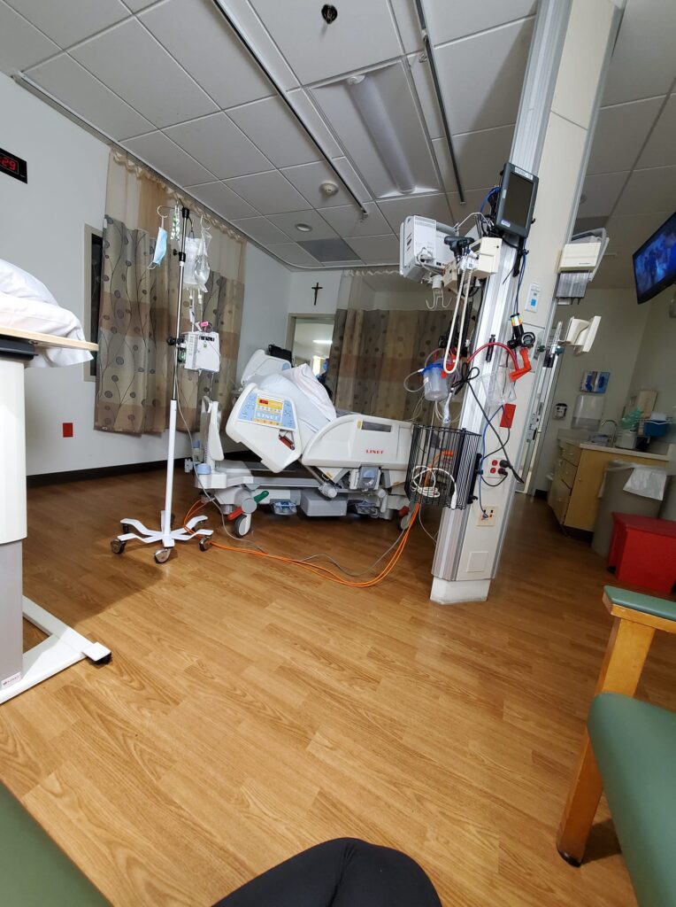 an ICU room with a hospital bed, an IV with wires and plugs. a window with open blinds in the corner and a covered window next to it