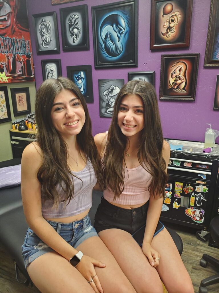 twin girls with long brown hair sitting with their arms around each other smiling at the camera in a room with a purple wall and paintings hung behind them