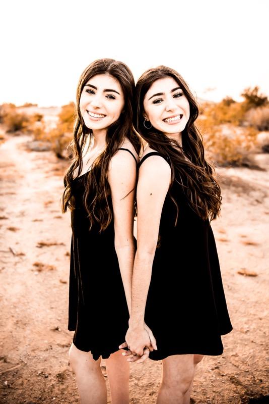 twin girls with long dark hair, standing back to back in the desert in black sleeveless dressings clasping hands and smiling at the camera